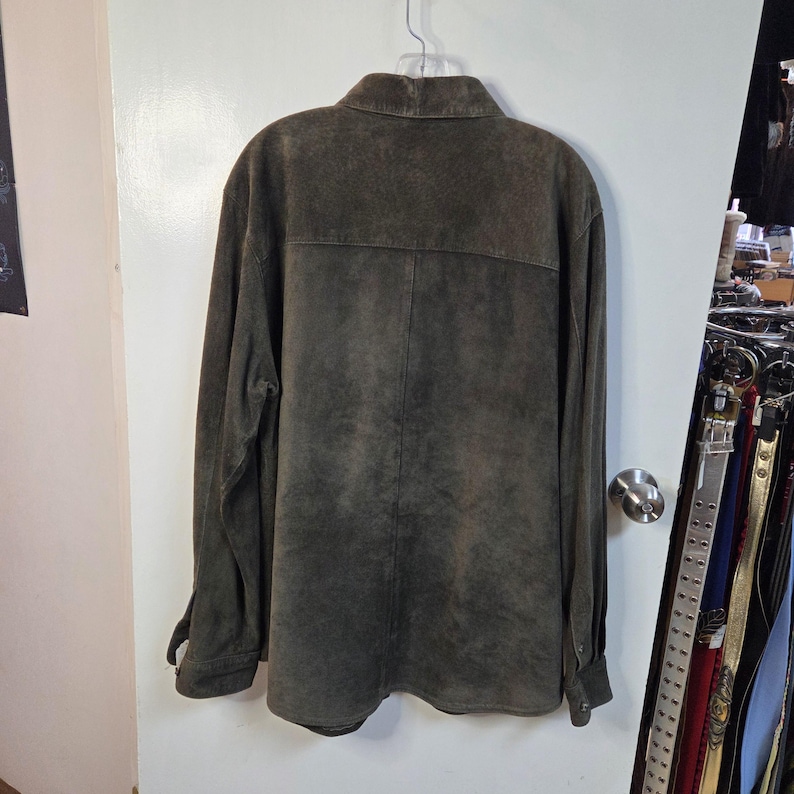 Pu&ograve; includere: Giacca a camicia in camoscio verde oliva scuro con maniche lunghe e polsini abbottonati. La giacca ha un colletto e una chiusura con bottoni sul davanti. La parte posteriore della giacca &egrave; visibile, appesa a una gruccia.