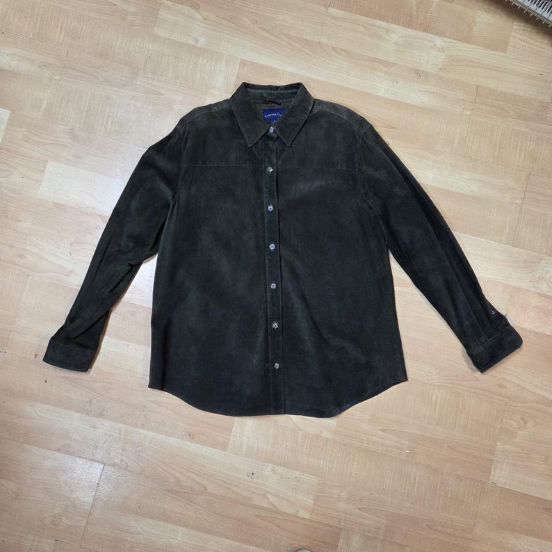 Pu&ograve; includere: Una camicia marrone scuro a maniche lunghe con colletto e bottoni. La camicia ha una consistenza simile alla pelle scamosciata e chiusure con bottoni. La camicia &egrave; stesa su una superficie di legno.
