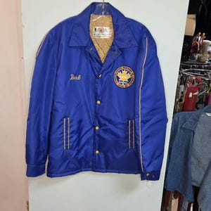Könnte beinhalten: Blaue Satinjacke mit goldfarbenen Zierleisten und Steppfutter. Die Jacke hat eine Knopfleiste, zwei Vordertaschen und einen Kragen. Auf der linken Brust steht der Name "Herb" in goldener Schrift, und ein Aufnäher mit Text und Logo befindet sich auf der rechten Brust.