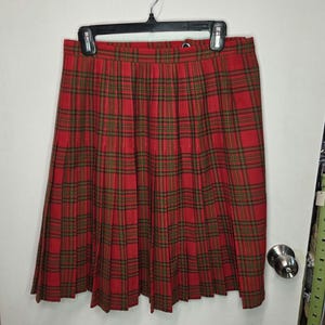 May include: A red tartan pleated skirt with green and black checks hangs on a black hanger. The skirt is made of a woven fabric. The skirt is a vintage style. The skirt is a classic tartan pattern.