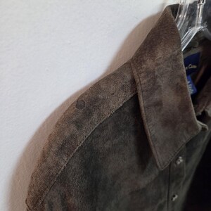 Pu&ograve; includere: Primo piano di una giacca in camoscio verde oliva scuro. Il colletto e i dettagli delle spalle sono a fuoco, mostrando le variazioni di consistenza e colore. La giacca &egrave; appesa a un'appendiabiti.