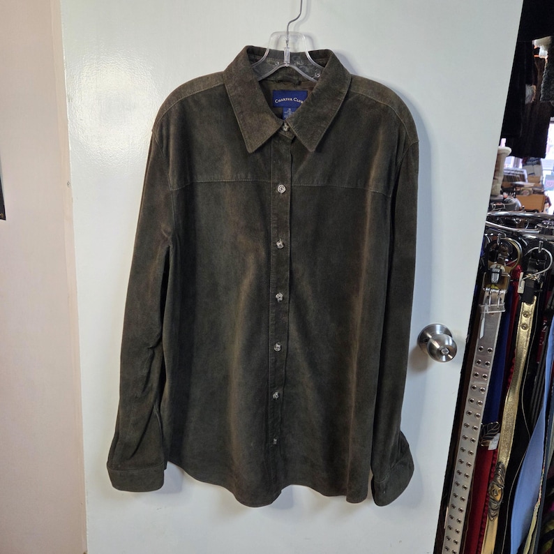 Pu&ograve; includere: Una camicia abbottonata a maniche lunghe di colore verde oliva tenue. La camicia ha un colletto ed &egrave; appesa a una gruccia di plastica trasparente. Il tessuto sembra essere camoscio o un materiale simile al camoscio. La camicia ha uno stile classico e casual.