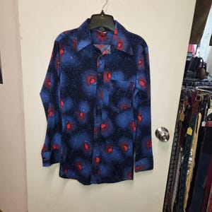 May include: A long-sleeve button-down shirt featuring a dark blue background with a speckled pattern of light blue and vibrant red circular shapes. The shirt includes a pointed collar, a single chest pocket, and buttoned cuffs on the sleeves.