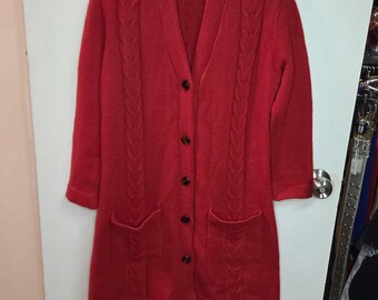 Può includere: Cardigan lungo rosso con scollo a V e chiusura con bottoni. Il maglione presenta dettagli a trecce sul davanti e sulle maniche, con due tasche anteriori. L'indumento è appeso a una gruccia di plastica trasparente.