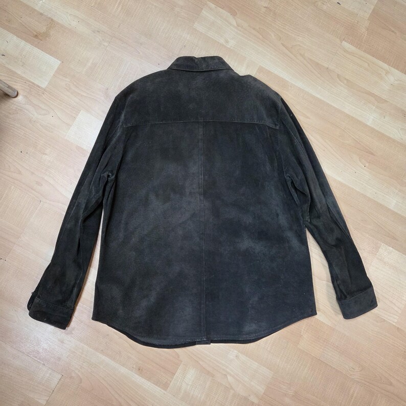 Pu&ograve; includere: Giacca in pelle scamosciata marrone scuro con maniche lunghe e polsini abbottonati. La giacca ha un colletto classico e una chiusura frontale con bottoni. La parte posteriore della giacca presenta un dettaglio a carr&egrave;. La giacca &egrave; appoggiata su una superficie di legno chiaro.