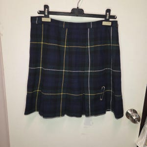 May include: A navy blue, green, and yellow tartan skirt hanging on a black hanger. The skirt features a pleated design and is secured with a safety pin. The skirt is made of a woven fabric.