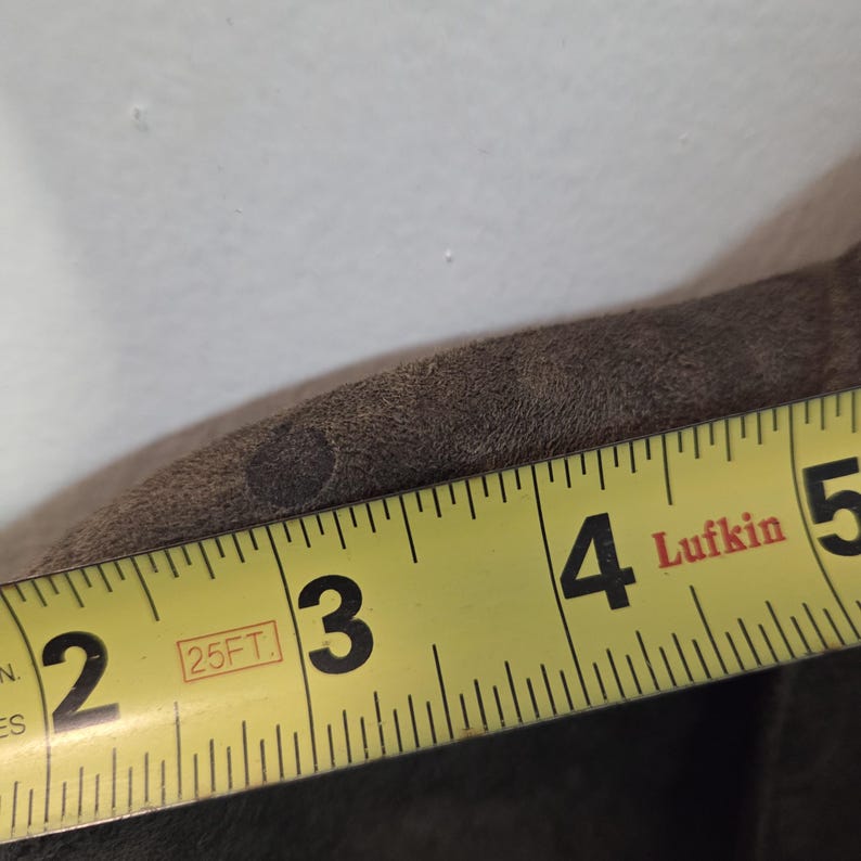 Pu&ograve; includere: Primo piano di un oggetto in pelle marrone con un metro a nastro giallo Lufkin. Il nastro mostra misure da 5 a 12,7 cm. La pelle ha una superficie strutturata e una piccola macchia scura.