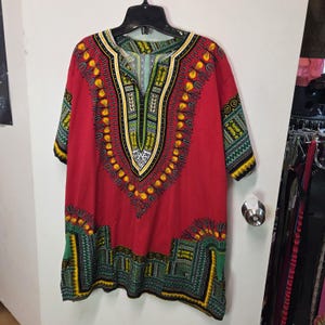 May include: A red dashiki shirt featuring a V-neckline and short sleeves. The garment is decorated with intricate geometric and floral patterns in yellow, green, black, and white, concentrated around the neckline, sleeves, and bottom hem.