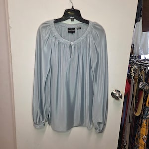 May include: A light blue long-sleeve blouse with a satin-like finish, featuring a round neckline with a small keyhole opening, a single button closure at the collar, and gathered fabric details. The interior label reads HALSTON X.