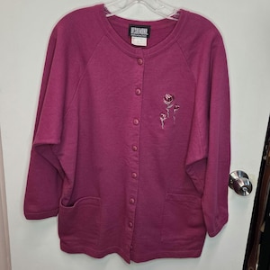 May include: A magenta button-up cardigan with three-quarter sleeves and two front pockets. The cardigan has a crew neck and a small embroidered floral design on the left side. The label reads "DESIRMOND."