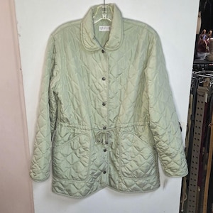 May include: A pale green quilted jacket with a round collar and button fastening. The jacket has a drawstring waist, two front pockets, and a diamond pattern. The jacket is hanging on a hanger.