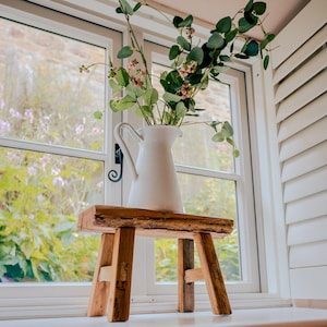 May include: A white ceramic jug with a handle is placed on a small, rustic wooden stool. The jug contains green and pink floral stems. The stool is in front of a window overlooking a garden.