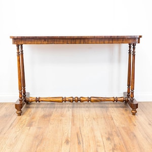 May include: A rectangular wooden console table with a dark brown finish and visible wood grain. The table features four slender, turned legs connected by a horizontal stretcher with decorative spindle detailing, resting on two flat base supports.