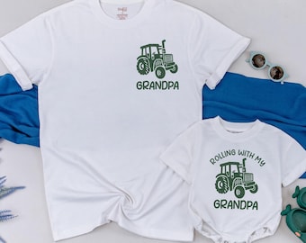 May include: Two white t-shirts, one adult and one infant, with green tractor graphics. The adult shirt says "GRANDPA" below the tractor. The infant shirt says "ROLLING WITH MY GRANDPA". Sunglasses and green shoes are also in the image.