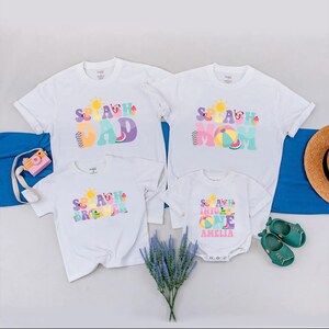 May include: Four white t-shirts with the word "Splash" in colourful designs. The shirts say "Dad", "Mom", "Brother", and "Into One Amelia". A pink toy camera, a straw hat, and green shoes are also in the image.