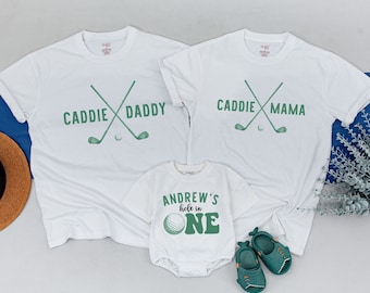 May include: White t-shirts with green golf club and ball graphics. The shirts say "Caddie Daddy" and "Caddie Mama". A white onesie with a green golf ball and the text "Andrew's hole in one".