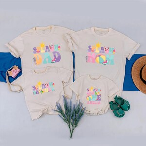 May include: A family set of beige t-shirts and a onesie with the text "Splash Dad", "Splash Mom", "Splash Brother", and "Hello Amelia". The shirts have colourful summer-themed graphics. A pink toy camera, a straw hat, and green shoes are also visible.