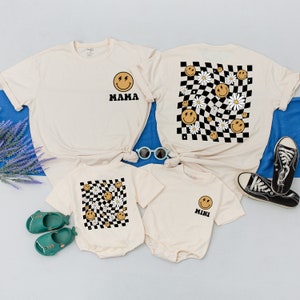 May include: Three cream-colored t-shirts with a black and white checkered pattern and smiley faces. The largest shirt says "MAMA" and the smallest shirt says "MINI".