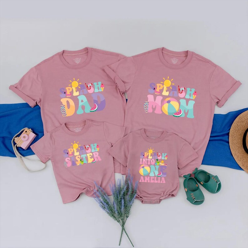 May include: Four matching mauve t-shirts and a onesie with the text "SPLASH" and family member titles. The shirts have colourful summer graphics. A camera, hat, and shoes are also in the image. Family vacation apparel.