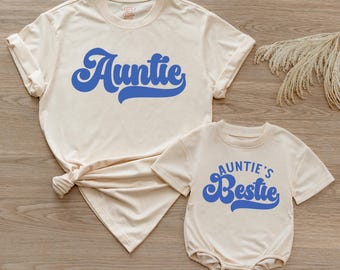 May include: A cream-colored short-sleeve t-shirt with the word Auntie printed in blue script and a matching cream-colored short-sleeve bodysuit with the text Auntie's Bestie printed in blue script, both featuring a rounded neckline.