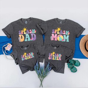 May include: Four dark grey t-shirts with the words "SPLASH DAD", "SPLASH MOM", "SPLASH BROTHER", and "SPLASH SISTER" in colourful, retro-style lettering. Each shirt features a sun graphic. The shirts are arranged with a blue cloth, a straw hat, and baby shoes.
