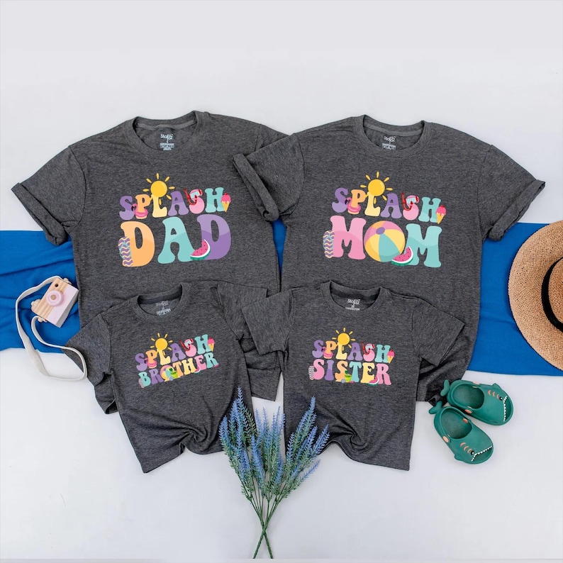 May include: Four dark grey t-shirts with the words "SPLASH DAD", "SPLASH MOM", "SPLASH BROTHER", and "SPLASH SISTER" in colourful, retro-style lettering. Each shirt features a sun graphic. The shirts are arranged with a blue cloth, a straw hat, and baby shoes.