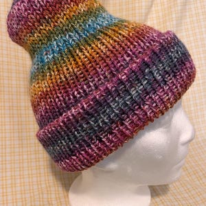 May include: A hand-knitted beanie featuring a ribbed texture and a folded brim. The yarn displays a variegated color pattern with blended shades of purple, blue, orange, yellow, and dark grey, creating a multi-colored effect throughout the fabric.