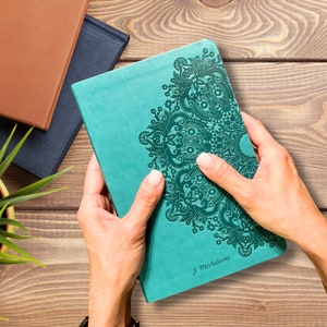 May include: Teal-colored journal with an embossed floral design on the cover. The journal has the name "J. McAdams" printed at the bottom. Other journals in brown and navy blue are visible in the background.