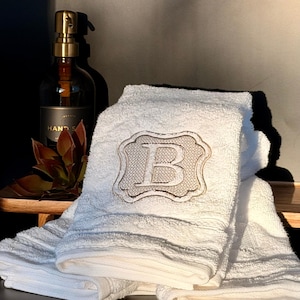 May include: White bath towel with an embroidered letter "B" in a decorative frame. A dark glass bottle with a gold pump dispenser is visible in the background, along with a small succulent plant. The towel is folded and stacked.