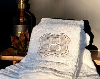 May include: A white terry cloth hand towel featuring a centered, embroidered beige patch with a scalloped border and the letter B in a serif font. The towel has a textured, ribbed pattern and is folded neatly in a stack.