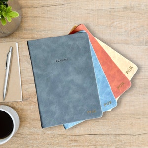 May include: Four leather-bound planners in grey, light blue, coral, and cream. The word "PLANNER" is embossed on the grey planner. Each planner has the initials "PGK" in gold lettering. The planners are arranged on a wooden surface.