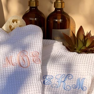 Puede incluir: Dos albornoces blancos de tejido de gofre con iniciales bordadas en rosa y azul. Las batas están dobladas y se exhiben con botellas de vidrio marrón con bombas doradas, una esponja vegetal y una planta suculenta, creando una estética de spa.