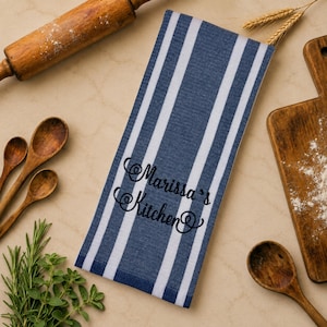 May include: A rectangular kitchen towel featuring a pattern of vertical navy blue and white stripes. The centre of the towel is embroidered with the black cursive text "Marissa's Kitchen" in a decorative script font with swirling flourishes.