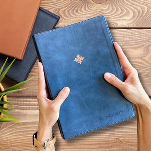 Könnte beinhalten: Ein blaues Lederjournal mit goldenem floralem Design und dem Namen "C. Nelson" auf dem Einband. Weitere Journale in Braun und Schwarz sind im Hintergrund zu sehen. Das Journal wird von einer Person gehalten.