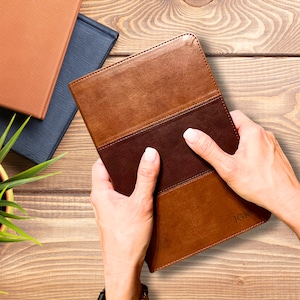 May include: A brown leather journal with a dark brown center panel and light brown border is held open by two hands. The initials "JGR" are embossed on the lower right corner. Other books are visible in the background.