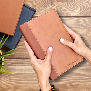 May include: A tan leather journal with a textured spine and the name "E. Christensen" embossed on the cover. The journal is held by hands, with two other journals in the background.