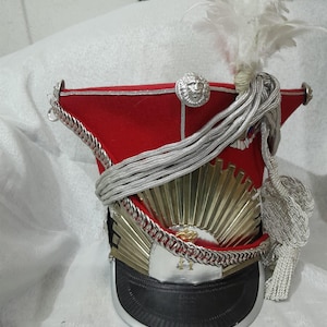 Könnte beinhalten: Ein roter Militärhut mit schwarzem Schirm, silbernen Akzenten und einem goldenen Sonnenstrahl-Design. Der Hut hat ein silbernes Löwenemblem, silberne Seil-Details und eine weiße Federquaste. Ein dekoratives Stück.