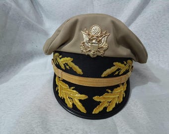 May include: Tan military hat with a gold emblem and gold embroidered leaf designs. The hat features a black brim with gold accents and a gold band. A classic military style.