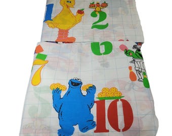 May include: A white bed sheet featuring a grid pattern and colorful Sesame Street characters. Big Bird is next to the number one, while Cookie Monster holds cookies near the number ten. Other numbers and characters are printed across the fabric.