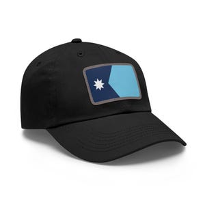 Op de afbeelding: Zwarte baseballpet met een rechthoekige patch. De patch heeft een ontwerp met een witte ster, marineblauwe en lichtblauwe secties en een grijze rand. De pet heeft een gebogen klep en een knoop aan de bovenkant.