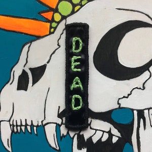 May include: A white skull with a black crescent moon and a green and yellow spiky mohawk. The skull has a black and green fabric patch that says "DEAD" on it.
