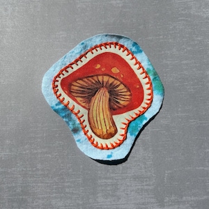 May include: A fabric patch featuring a red mushroom with white spots and a brown stem. The mushroom is set against a blue and white tie-dye background, with orange stitching around the edge.