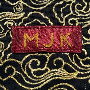 Może przedstawiać: Czerwona metka z tkaniny z wyhaftowanymi złotymi nićmi literami "MJK".