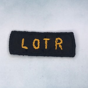 Puede incluir: Un parche rectangular negro con las letras "LOTR" bordadas en dorado. El parche tiene un borde texturizado y es probablemente un artículo de colección o decorativo.