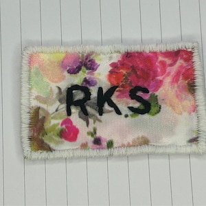 May include: A white fabric patch with a floral print and the letters "RKS" embroidered in black.