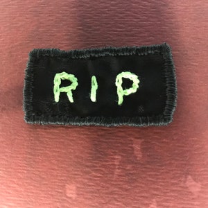 May include: A black rectangular patch with the letters "RIP" embroidered in light green thread. The patch has a black border and is set against a reddish-brown background.