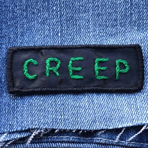 Op de afbeelding: Een zwarte patch met groene geborduurde letters die "CREEP" lezen op een blauwe denim achtergrond.