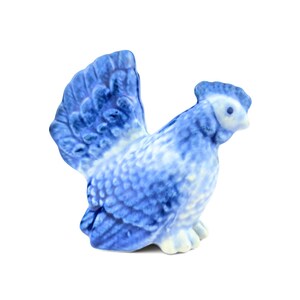 May include: A small ceramic figurine of a hen with a blue and white mottled pattern. The bird is depicted in a seated position with its tail feathers fanned upward, featuring detailed molded textures on the wings and body, and a small comb.
