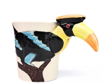 May include: A white ceramic mug with a painted image of a blue and black bird with a yellow beak. The bird's head is the handle of the mug.