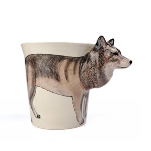 May include: A ceramic coffee mug with a wolf design. The wolf's head is 3D and extends from the side of the mug. The wolf is brown and gray with a white background.